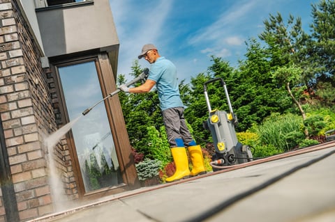A man in yellow boots uses a pressure washer to clean the exterior walls of a home