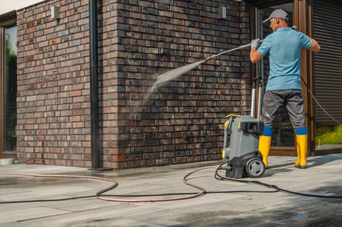 A man in yellow boots and gloves operates a pressure washer, cleaning a brick wall of a contemporary house while standing on a paved surface under clear skies.