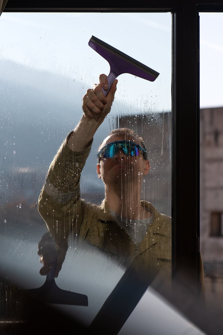 Worker performing exterior window cleaning using professional squeegee tool. Natural daylight, realistic urban setting. Concept of property maintenance, hygiene standards and service industry operations.