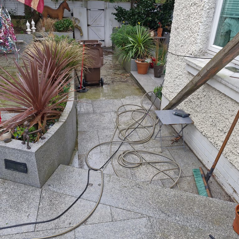 Person pressure washing a residential patio surrounded by potted plants, equipment, and an American flag