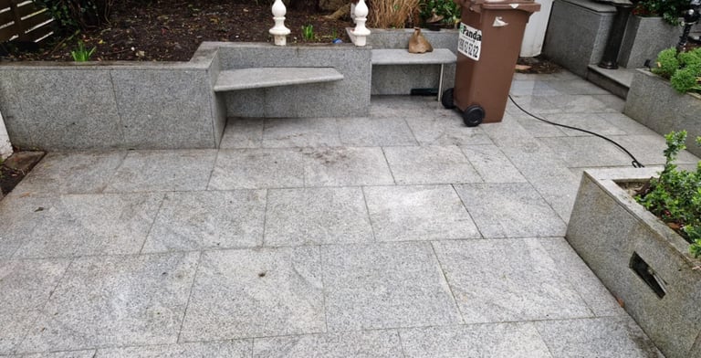 Gray paved patio area with concrete bench seating and planters, surrounded by green vegetation and a brown trash bin