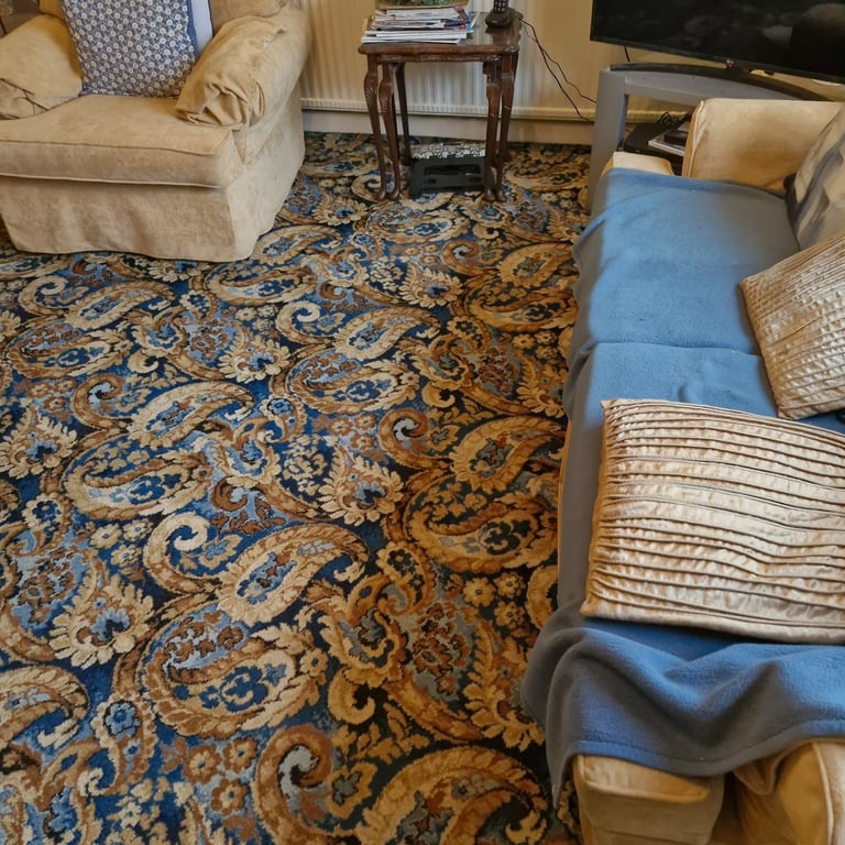 Living room with ornate blue and gold patterned rug, beige armchairs, wooden side table, and blue upholstered ottoman