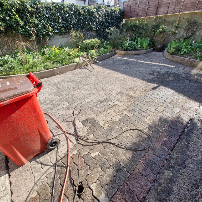 Red wheelie bin and garden hose on a brick paved backyard patio with raised garden beds and ivy-covered fence