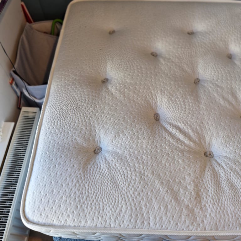 White quilted mattress with button tufting and gray border, displayed indoors with storage items nearby